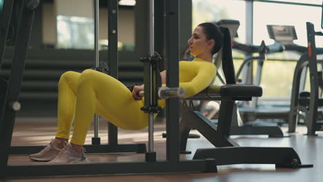 A-young-woman-in-a-yellow-tracksuit-lifts-her-body-with-a-barbell-to-train-the-back-of-her-thigh-and-buttocks.-Weight-training-in-the-smith-simulator.-Lifting-the-waist-and-body-with-weight-in-the-gym